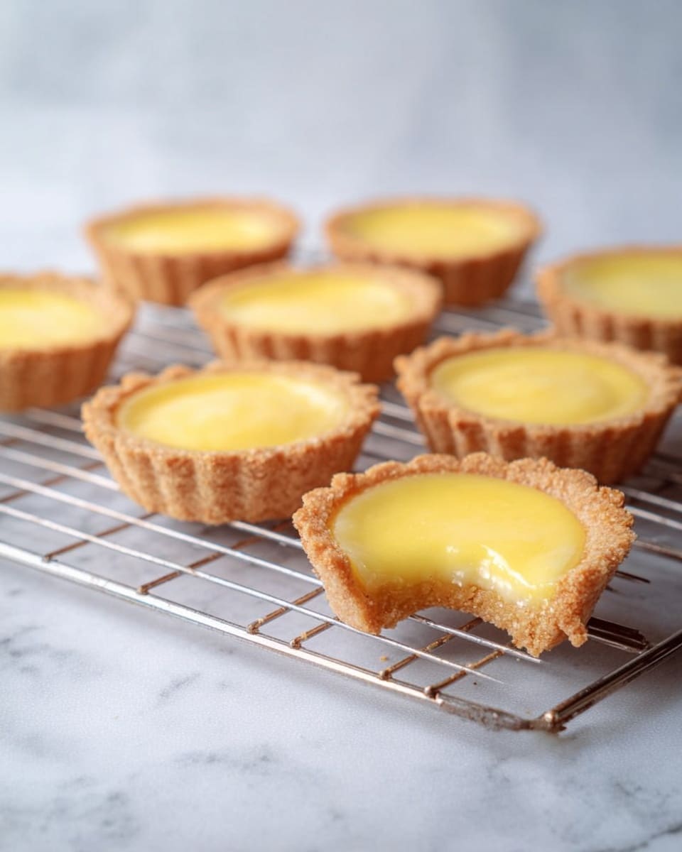 The image shows eight small tartlets with a golden-yellow custard filling and light brown crumbly crusts arranged on a silver cooling rack over a white marbled surface. One tartlet in the front has a small bite taken from its edge, revealing the smooth, creamy interior and the crumbly texture of the crust. The tartlets have a fluted edge and a shiny, smooth custard that fills each crust evenly. The background is soft and light, with the focus on the tarts in the front while those further back are slightly blurred. photo taken with an iphone --ar 4:5 --v 7