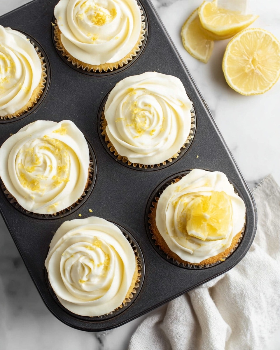 A dark metal cupcake tray holds four cupcakes, each with two visible layers: a light yellow cake base and a thick, creamy white frosting on top shaped like a rose with soft, swirled petals. One cupcake in the lower right corner is decorated with a small bright yellow lemon wedge and light lemon zest sprinkled over the frosting. The tray is set on a white marbled surface, where two lemon wedges rest nearby along with a soft white cloth partially visible in the bottom corner. photo taken with an iphone --ar 4:5 --v 7