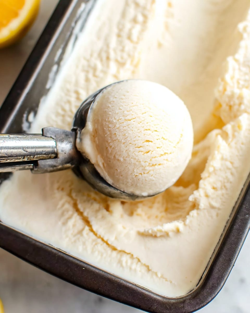 A close-up view of a shiny metal ice cream scooper holding a smooth, round scoop of creamy light beige ice cream resting inside a dark, rectangular baking pan filled with evenly spread ice cream. The ice cream has a soft, dense texture with slight scoop marks around the edges, and the background features a white marbled surface with a faint lemon shape blurred out on the side. photo taken with an iphone --ar 4:5 --v 7