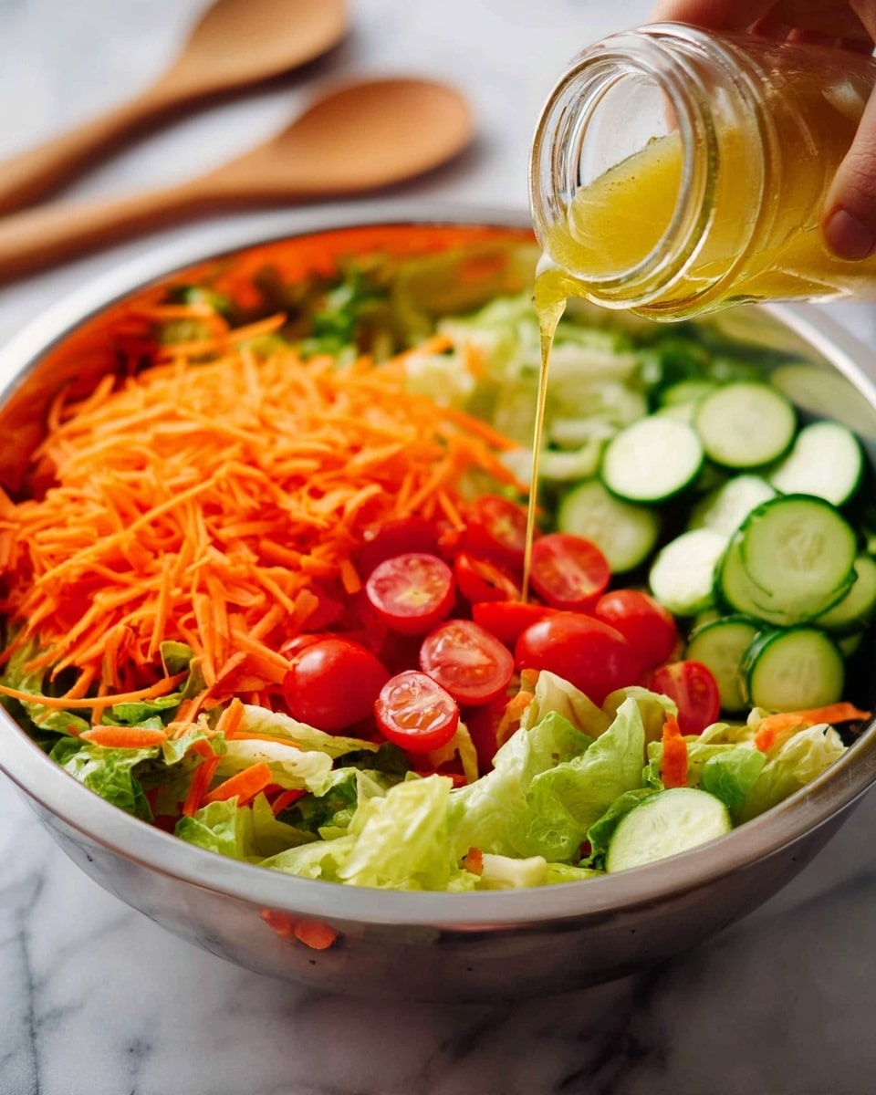 A large metal bowl filled with fresh salad divided into layers: bright green lettuce leaves at the bottom left, thin orange shredded carrots piled on top of the lettuce to the left, halved shiny red grape tomatoes forming a cluster in the middle, and several round, smooth light green cucumber slices stacked neatly to the right. A woman's hand is pouring a golden-yellow dressing from a small clear glass jar over the carrots. The bowl sits on a white marbled surface with two wooden spoons blurred in the background. Photo taken with an iphone --ar 4:5 --v 7
