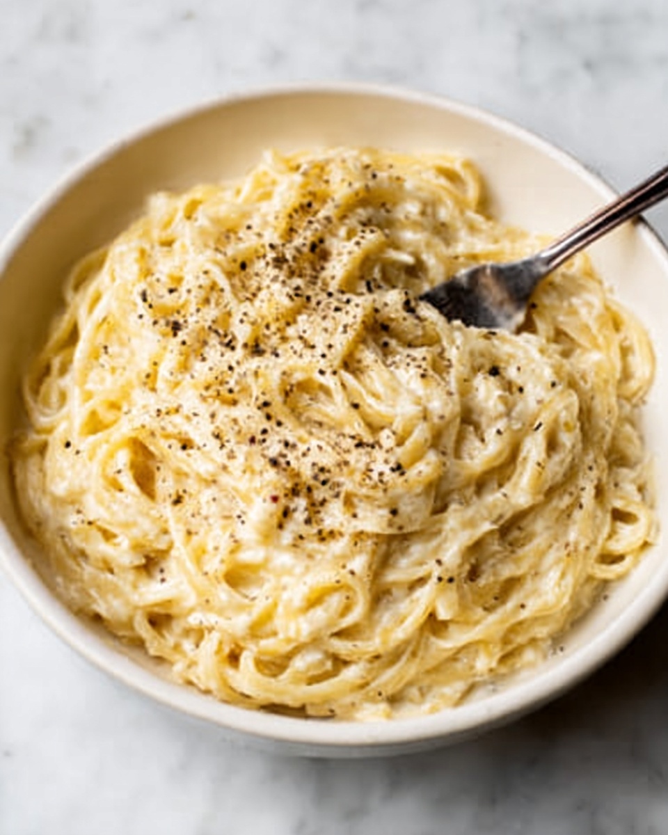 A white bowl filled with creamy fettuccine alfredo pasta, with thick sauce covering every strand. The pasta is pale yellow and looks soft, sprinkled evenly with black pepper on top. A fork rests inside the bowl, partially mixed into the noodles. The bowl sits on a white marbled surface that contrasts with the warm color of the pasta. photo taken with an iphone --ar 4:5 --v 7