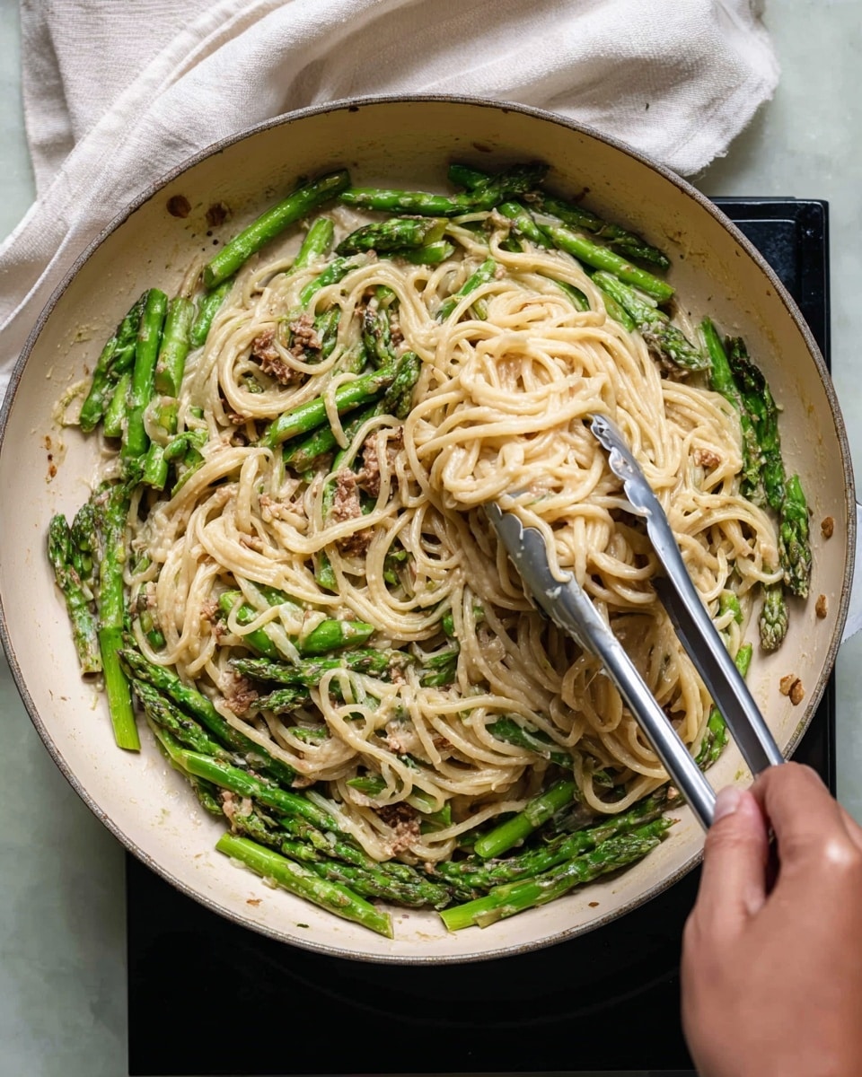 A close-up image shows a white pan filled with three main layers: at the bottom, there are bright green asparagus pieces evenly placed around the edge; in the center, a thick layer of creamy light beige pasta noodles is mixed with the asparagus; and scattered lightly throughout, small bits of browned protein add texture. A pair of metal tongs held by a woman's hand lifts a twisted bundle of pasta from the pan, showing slight steam and a rich sauce coating the noodles and vegetables. The pan sits on a black cooking surface set on a white marbled texture surface with a white cloth partially visible beside it. Photo taken with an iphone --ar 4:5 --v 7