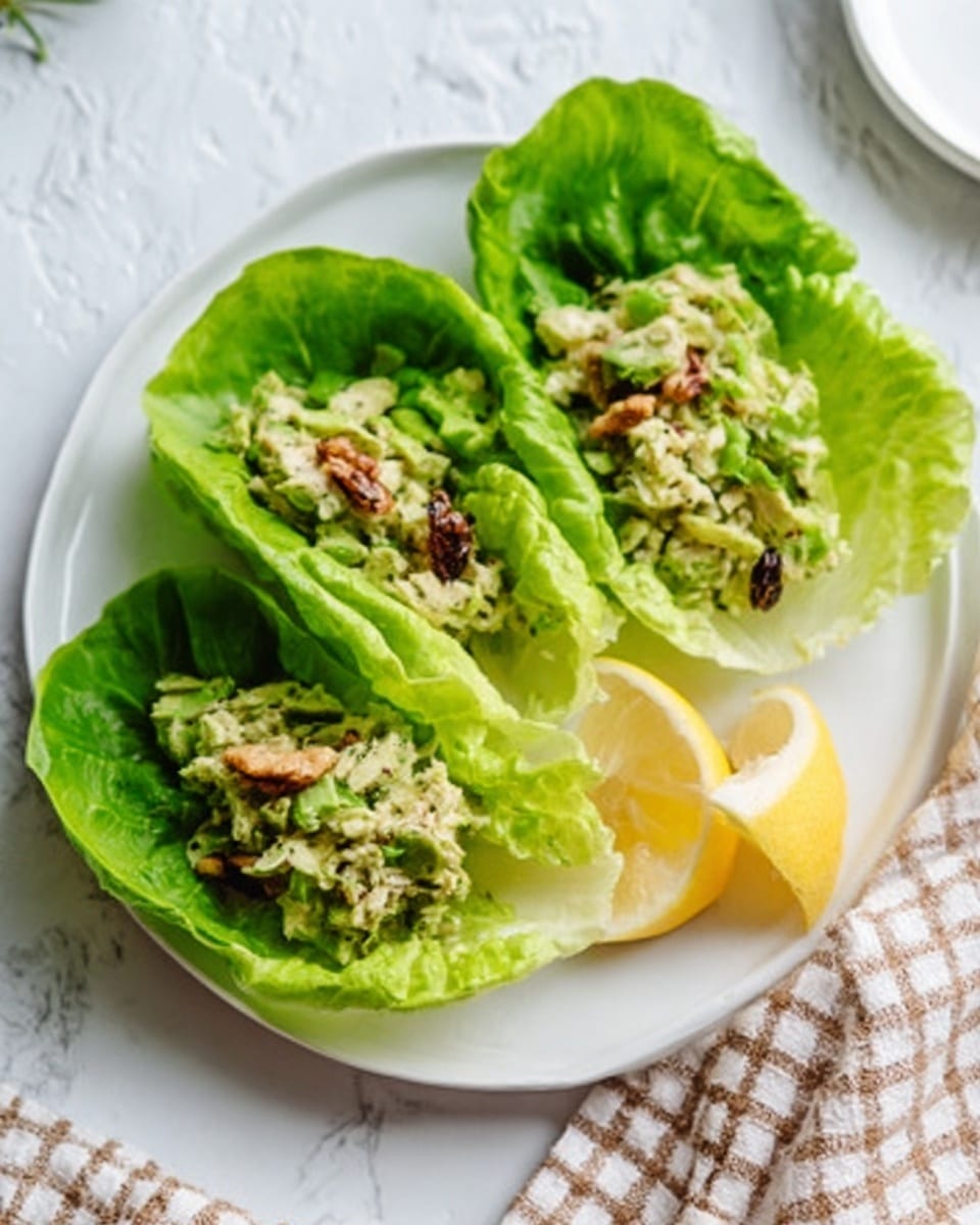 The image shows three lettuce wraps placed side by side on a round white plate. Each wrap is made of bright green, crisp lettuce leaves that form a bowl-like shape holding a filling inside. The filling appears chunky with small pale green pieces mixed with bits of darker ingredients that look like nuts or dried fruits. A lemon wedge is placed on the right side of the plate, and the plate is set on a white marbled surface with a white round plate and a checkered beige cloth nearby. photo taken with an iphone --ar 4:5 --v 7