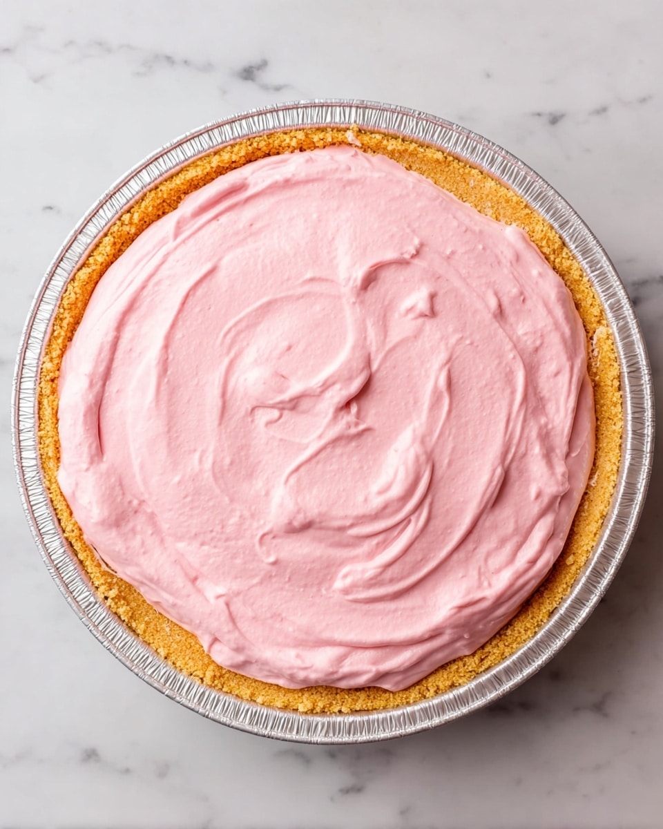 The image shows a pie in a silver foil pie dish placed on a white marbled surface. The pie has two layers: the bottom layer is a golden brown crumb crust with a slightly rough texture, fitting snugly in the pie dish. The top layer is a smooth, thick pink cream spread evenly across the crust with soft swirls visible on its surface. The pie looks fresh and creamy with the pink layer being the most colorful part against the pale crust and foil dish. photo taken with an iphone --ar 4:5 --v 7