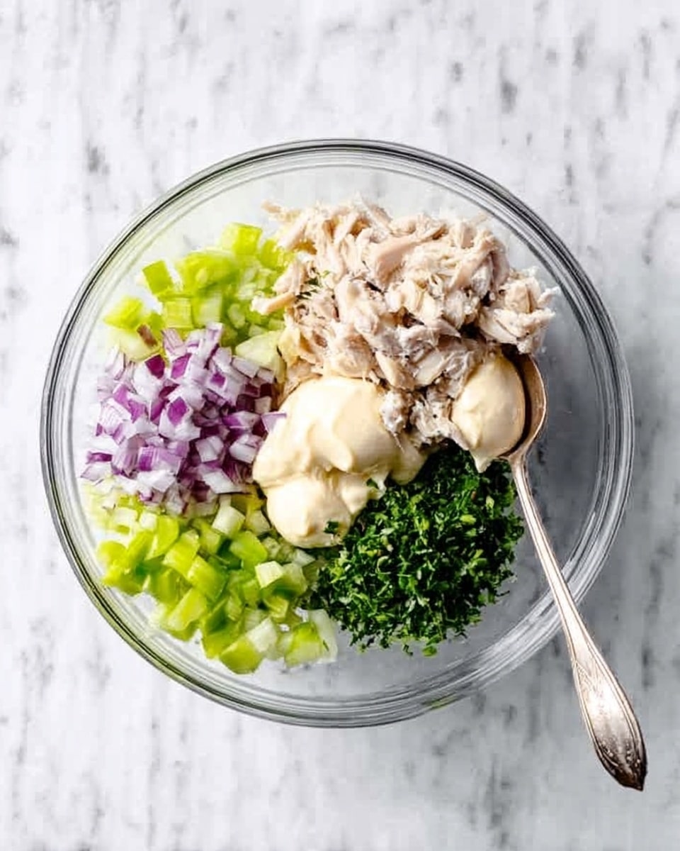 A clear glass bowl is placed on a white marbled surface, filled with six distinct ingredients arranged in separate sections. There are small, light green celery pieces on the left bottom side, finely chopped purple-red onions on the left top, a small pile of light beige chicken pieces in the middle bottom, finely chopped green herbs on the right top, a spoonful of creamy white mayonnaise on the top center, and dollops of a pale beige mustard-like sauce near the center. A silver spoon lies on the white marbled surface to the right of the bowl, and a woman’s hand is holding the bowl from the top left side. The overall scene looks fresh, colorful, and clean. photo taken with an iphone --ar 4:5 --v 7