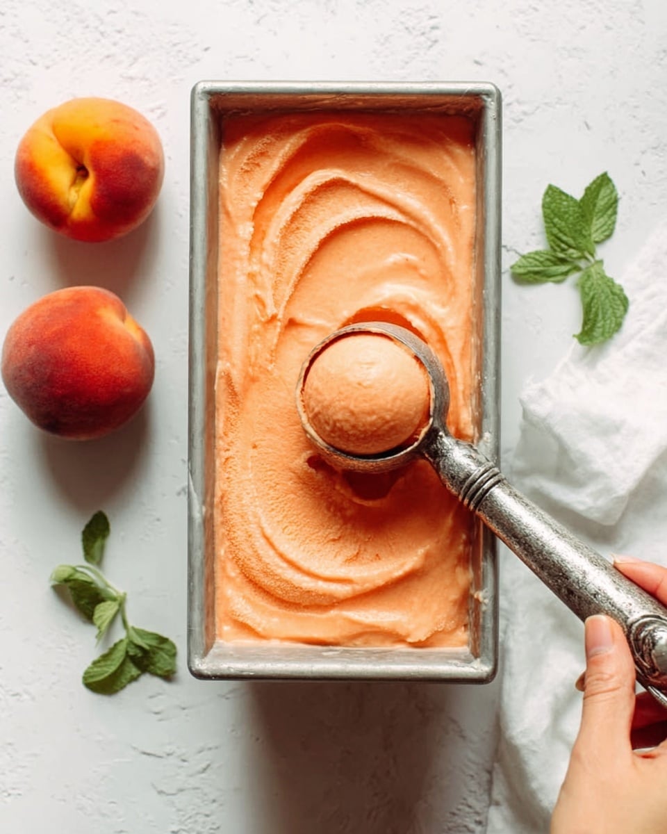The image shows a metal loaf pan filled with smooth, creamy peach-colored ice cream. A vintage ice cream scoop held by a woman's hand is lifting a round scoop out of the ice cream, creating soft, swirled textures in the surface. The background features a white marbled texture with two whole peaches and small green leaves scattered around, adding a fresh and natural look to the scene. The lighting is soft and natural, highlighting the rich texture and color of the ice cream. photo taken with an iphone --ar 4:5 --v 7