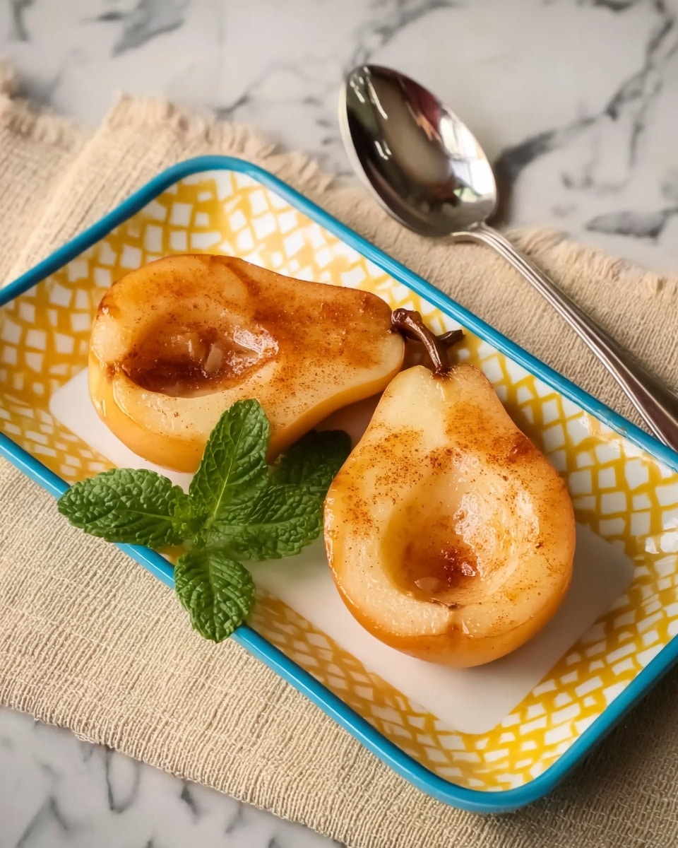 Two halves of a baked pear with a warm golden-brown color and soft texture rest on a white rectangular plate with a yellow diamond pattern and blue rim. The pear halves show a small scoop-shaped hollow in the center where the core was, sprinkled lightly with cinnamon. A small bunch of fresh green mint leaves lies between the pear halves. The plate is set on a white marbled surface with a beige textured napkin and a silver spoon nearby. Photo taken with an iphone --ar 4:5 --v 7