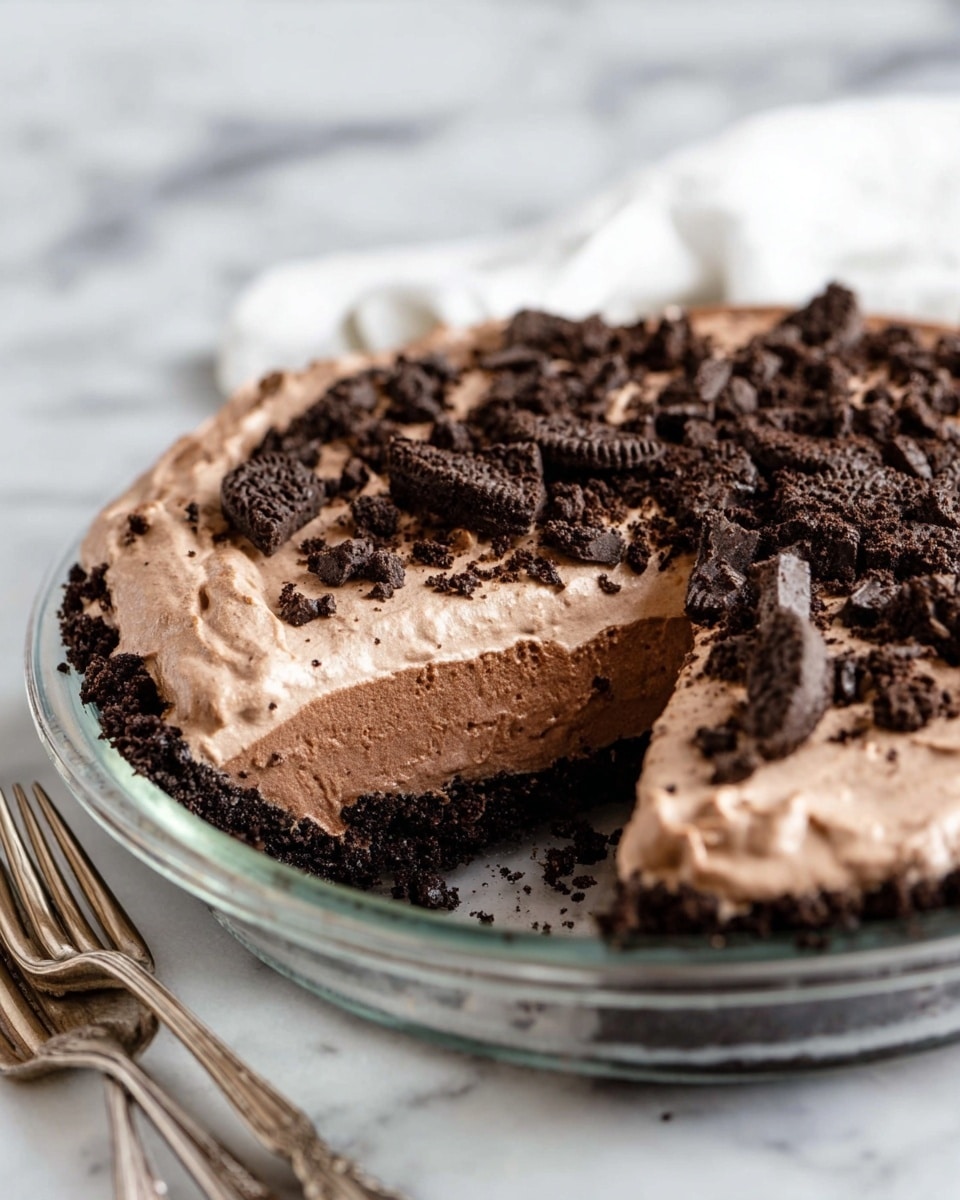 The image shows a pie with three main visible layers: the bottom crust is dark and crumbly, the middle filling is a lighter, smooth chocolate cream, and the top layer is a thicker, fluffy chocolate mousse. The pie is topped with uneven chunks of dark chocolate cookie pieces scattered over the mousse. The pie is in a clear glass pie dish sitting on a white marbled surface. In the foreground, two vintage-styled silver forks lie near the pie. Photo taken with an iphone --ar 4:5 --v 7