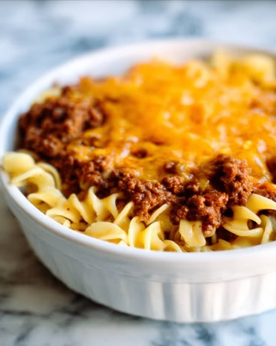 A close-up view of a white bowl filled with three layers: the bottom layer is curly egg noodles with a soft yellow color, the middle layer consists of brown cooked ground beef with a crumbly texture, and the top layer is melted cheddar cheese that is bright orange and smooth, covering the beef evenly. In the blurry background, there is a white marbled surface with a woman's hand slightly holding the bowl. The photo taken with an iphone --ar 4:5 --v 7