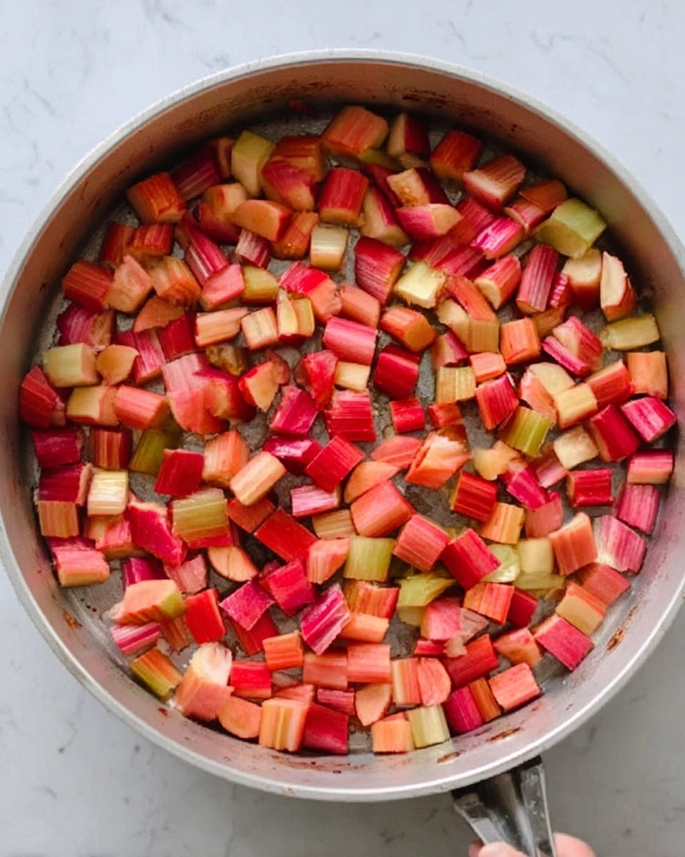 The image shows a white metal pan filled with many small pieces of colorful vegetables. The vegetables are chopped into small cubes and chunks with mostly red, orange, and cream colors from different kinds of root vegetables. There are also light green pieces that add more color. The vegetables fill the pan evenly, showing a mix of soft and firm textures. The pan rests on a white marbled surface, and a woman's hand is holding the handle from the right side. The photo taken with an iphone --ar 4:5 --v 7