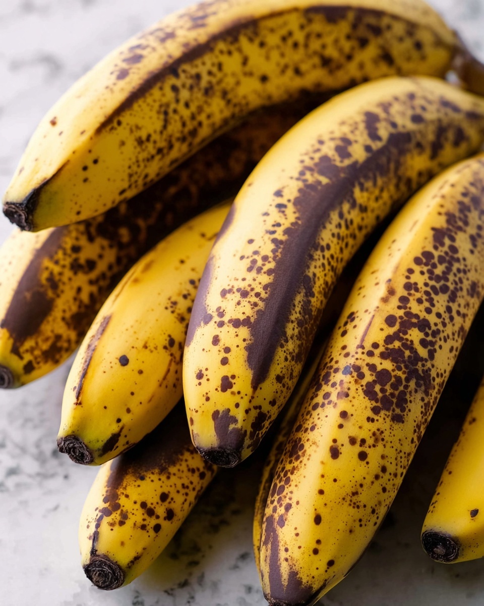 Raspberry Dark Chocolate Banana Bread Recipe 4 A bunch of ripe yellow bananas with many brown spots cover the peel, lying on a white marbled surface. The bananas are closely packed together, showing their dark brown stems at the top and smooth, curved bodies, with some bananas partly overlapping others. The color contrast between the bright yellow skin and the dark brown spots gives a natural, textured look to the fruit. Photo taken with an iphone --ar 4:5 --v 7