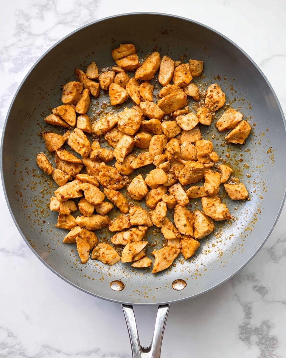 A gray frying pan with a shiny metal handle holds small, browned pieces of cooked chicken, scattered evenly across the bottom. The chicken pieces are golden brown with some slight darker spots from cooking, showing a textured surface with a slightly oily sheen. The pan sits on a white marbled surface with no other items around. Photo taken with an iphone --ar 4:5 --v 7