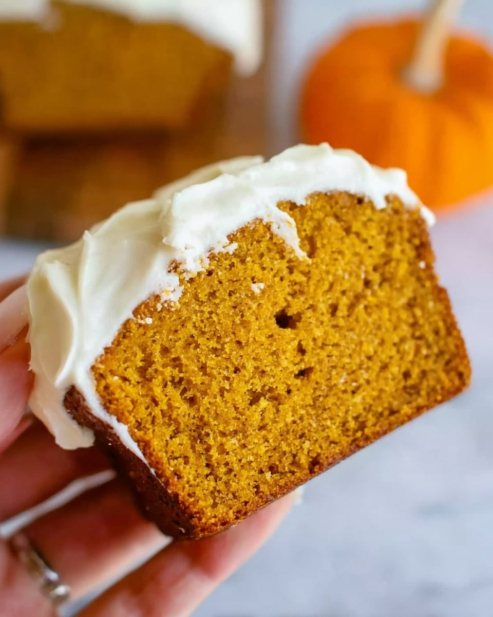 A close-up image showing a woman's hand holding a slice of pumpkin loaf with white frosting spread evenly on top. The loaf is golden brown with a slightly crumbly texture, and the frosting looks smooth and creamy. The background is softly blurred with an orange pumpkin shape visible, sitting on a white marbled surface. Photo taken with an iphone --ar 4:5 --v 7