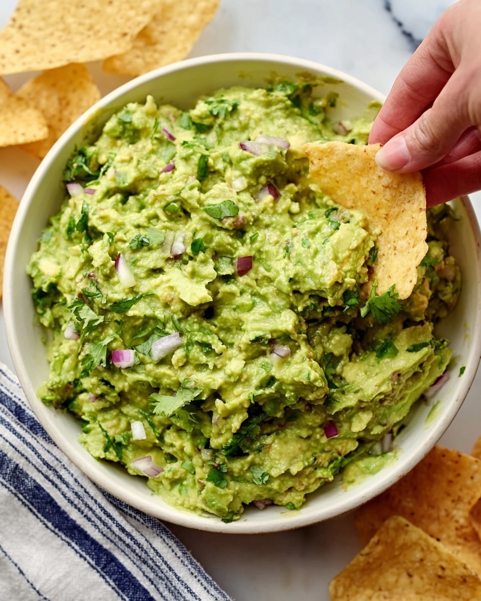 A close-up image of a white bowl filled with chunky green guacamole mixed with small pieces of red onion and cilantro. The guacamole has a slightly creamy but thick texture with visible bits of avocado. Around the bowl, there are light golden tortilla chips, with one chip being dipped into the guacamole by a woman’s hand. The bowl is placed on a white marbled surface with a striped cloth partially visible underneath. photo taken with an iphone --ar 4:5 --v 7