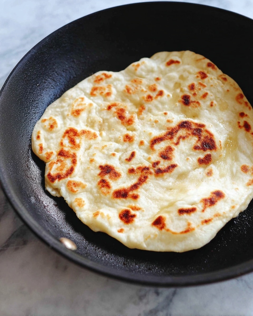 A single flatbread with a light cream color and browned spots is cooking in a black skillet. The flatbread has an uneven, rounded shape with soft textures and visible bubbles on its surface. The skillet sits on a white marbled surface. Photo taken with an iphone --ar 4:5 --v 7