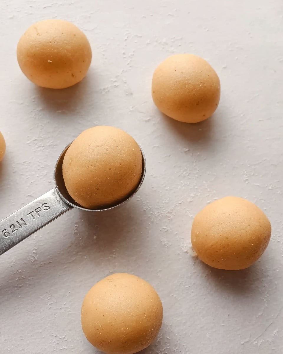 The image shows six smooth, light orange dough balls placed on a white marbled surface. One dough ball is in a measuring spoon labeled