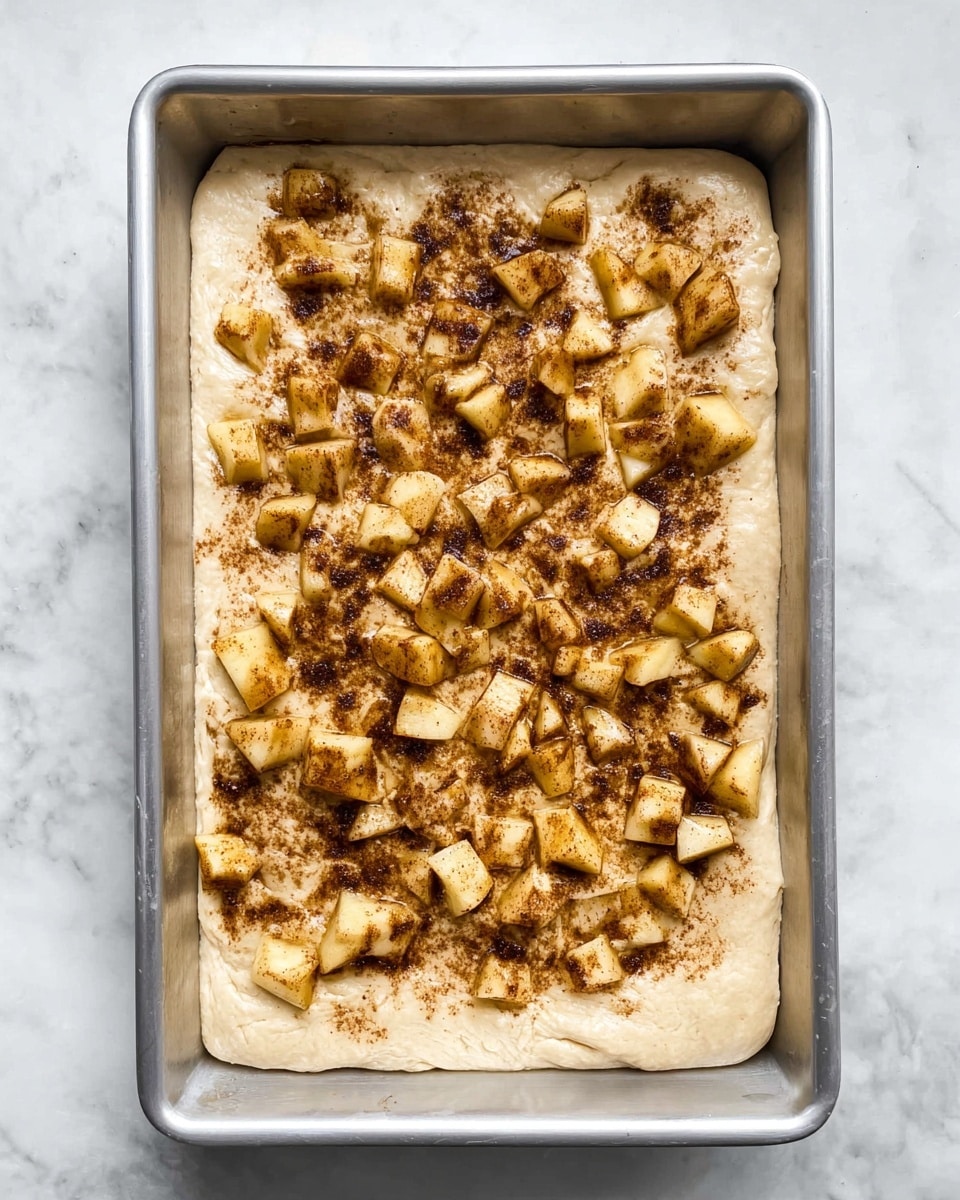 The image shows a rectangular metal baking pan filled with an even layer of light beige dough. On top of the dough, there are irregular pieces of light yellow chopped fruit with a brown cinnamon-like spice sprinkled over them, creating a speckled dark brown pattern against the lighter dough. The pan sits on a white marbled surface with a subtle veined pattern. The edges of the dough are slightly pushed up against the sides of the pan, and the fruit pieces are scattered mostly in the center, leaving some dough visible around the edges. photo taken with an iphone --ar 4:5 --v 7