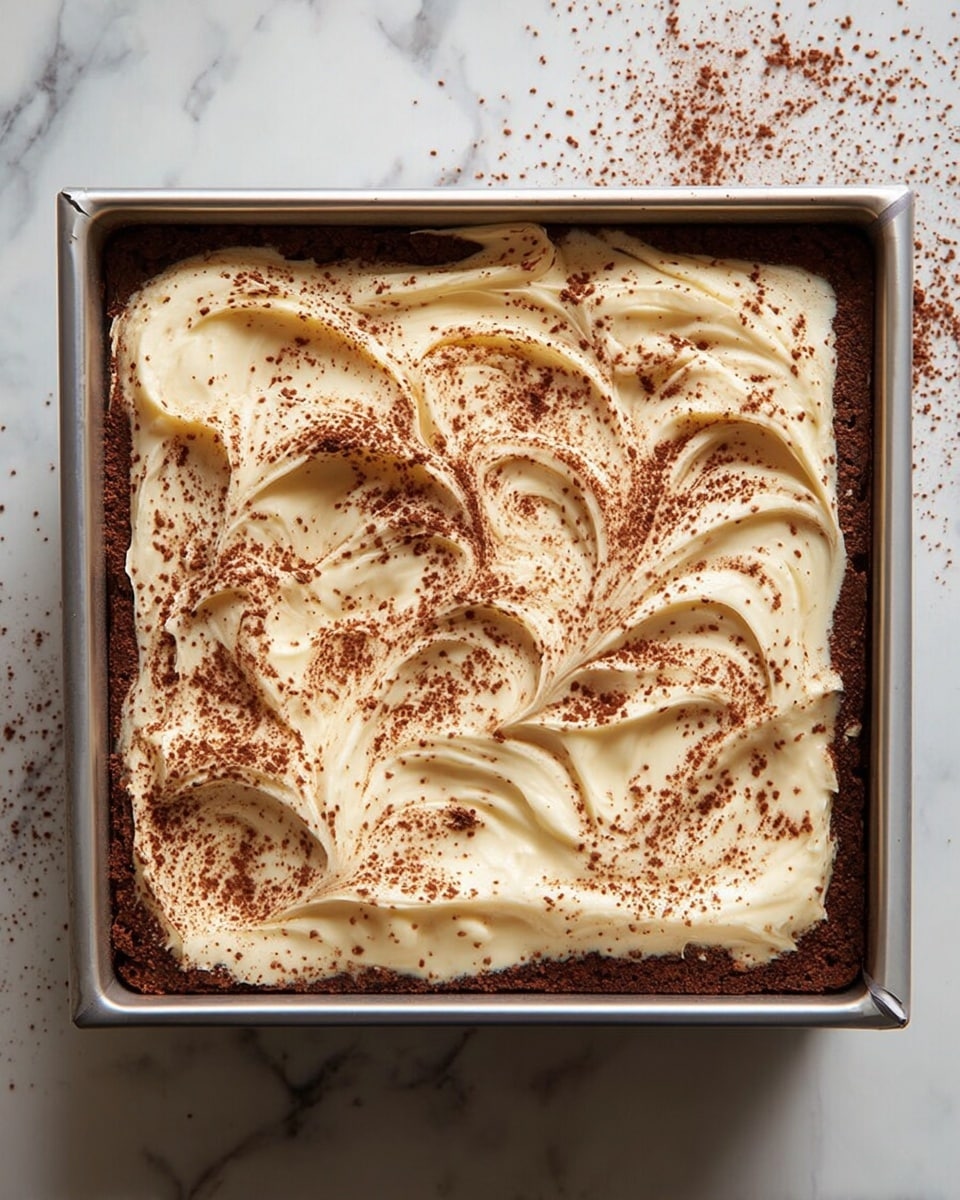 Limoncello Tiramisu Recipe 4 A square metal pan holds a layered dessert with a base layer of dark brown cake or cookie, topped by a thick layer of smooth, creamy white filling. The creamy layer has swirled, wavy peaks that add texture, and it is sprinkled lightly with a fine layer of cocoa powder, creating a mix of light cream and dark brown colors on top. The pan sits on a white marbled surface with some scattered cocoa powder around. Photo taken with an iphone --ar 4:5 --v 7