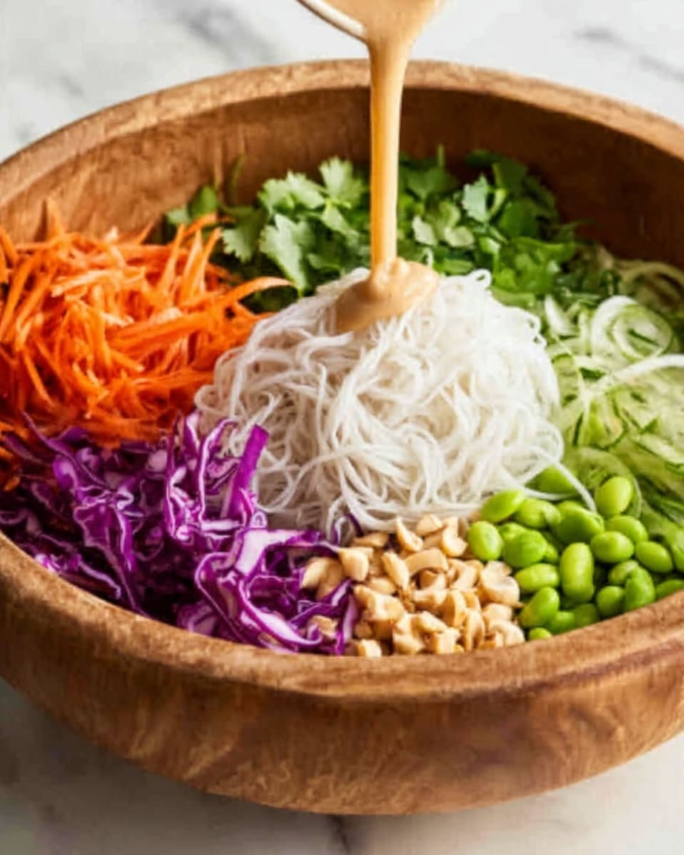 The image shows a wooden bowl filled with a colorful salad. The salad has three main layers visible: a bottom layer of thin, light beige noodles, a middle layer of shredded purple cabbage and thin orange carrot strips, and a top layer of green edamame beans and fresh cilantro leaves scattered around. On the top left side of the bowl, two bright green lime wedges rest on the salad. Small white sesame seeds are sprinkled evenly over the salad. Two wooden serving spoons are placed in the bowl, sticking out upwards. The bowl is set on a white marbled surface with a beige cloth next to it. photo taken with an iphone --ar 4:5 --v 7