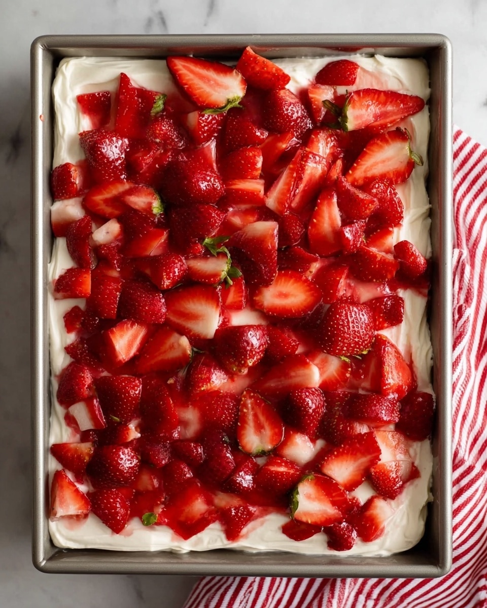 This image shows a square metal pan with two layers visible: the bottom layer is thick and creamy white, spread evenly with a smooth and soft texture, while the top layer is made up of many bright red strawberries, some whole with green tops and others sliced, covering the whole surface in a slightly shiny, fresh look. The pan is placed on a white marbled surface with a striped red and white cloth partly visible on the right side. Photo taken with an iphone --ar 4:5 --v 7