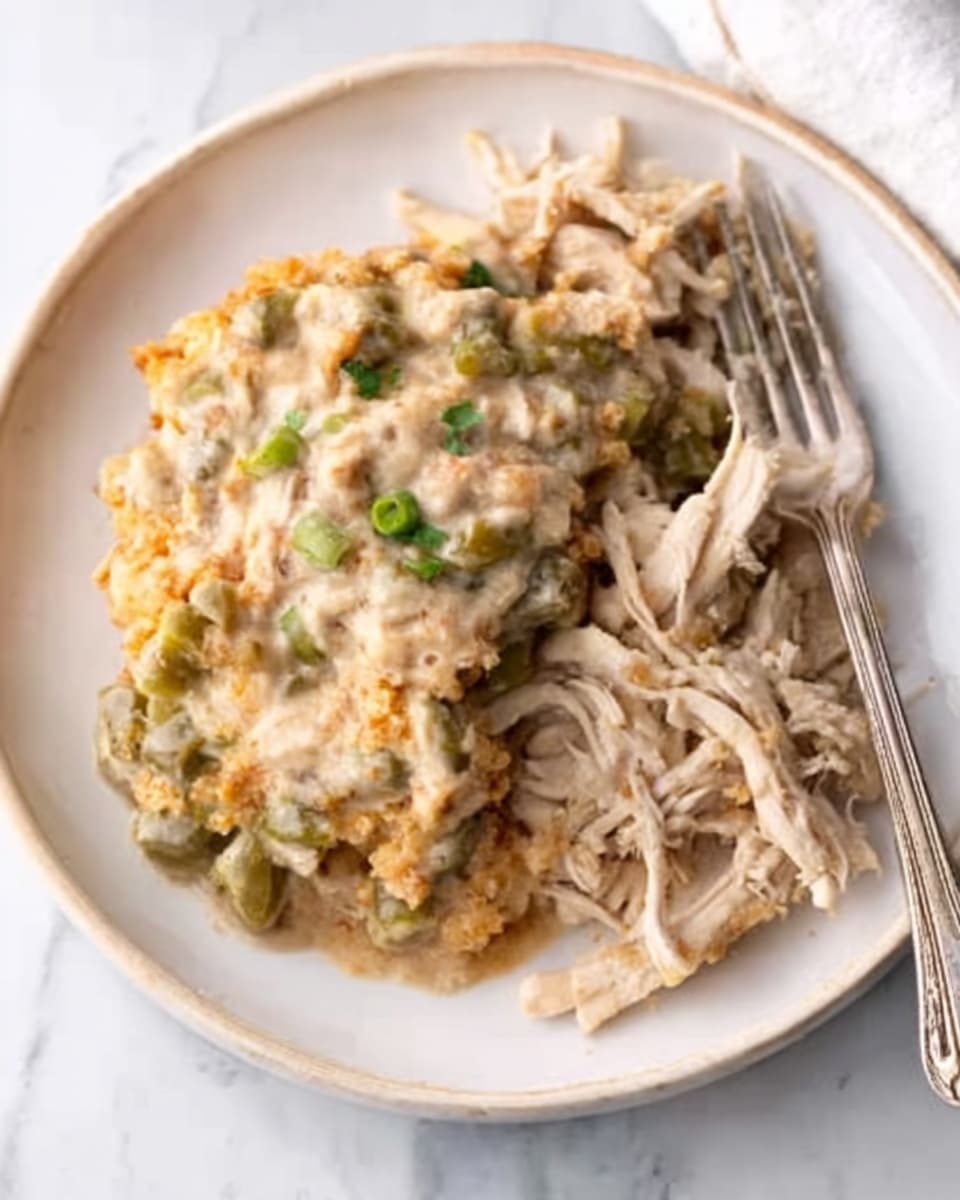 The image shows a close-up of a white plate on a white marbled surface, filled with a creamy mixture of green beans and a crunchy, golden-brown topping. Next to the green bean casserole, there is a portion of shredded light brown chicken meat. A fork is placed on the right side of the plate, and a woman's hand is holding the fork, ready to eat. The overall look is warm and homey, with a slight focus on the texture of the casserole and the tender chicken. photo taken with an iphone --ar 4:5 --v 7