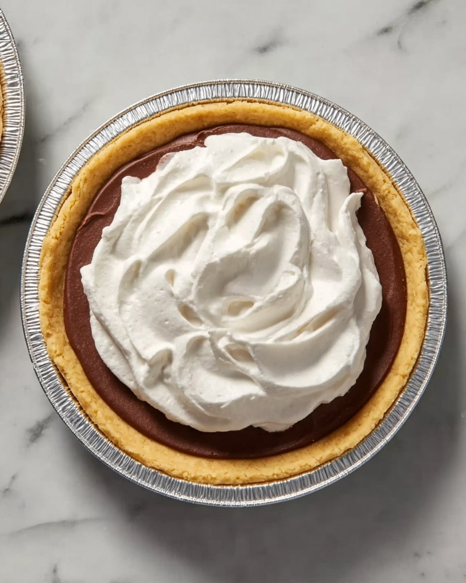 The image shows a single pie with three visible layers from bottom to top: a light golden brown crust that looks firm and thick, a smooth dark chocolate filling that fills most of the crust, and a thick layer of white fluffy whipped cream on top with swirling texture and soft peaks. The pie is in a silver foil pie tin, placed on a white marbled surface. Photo taken with an iphone --ar 4:5 --v 7