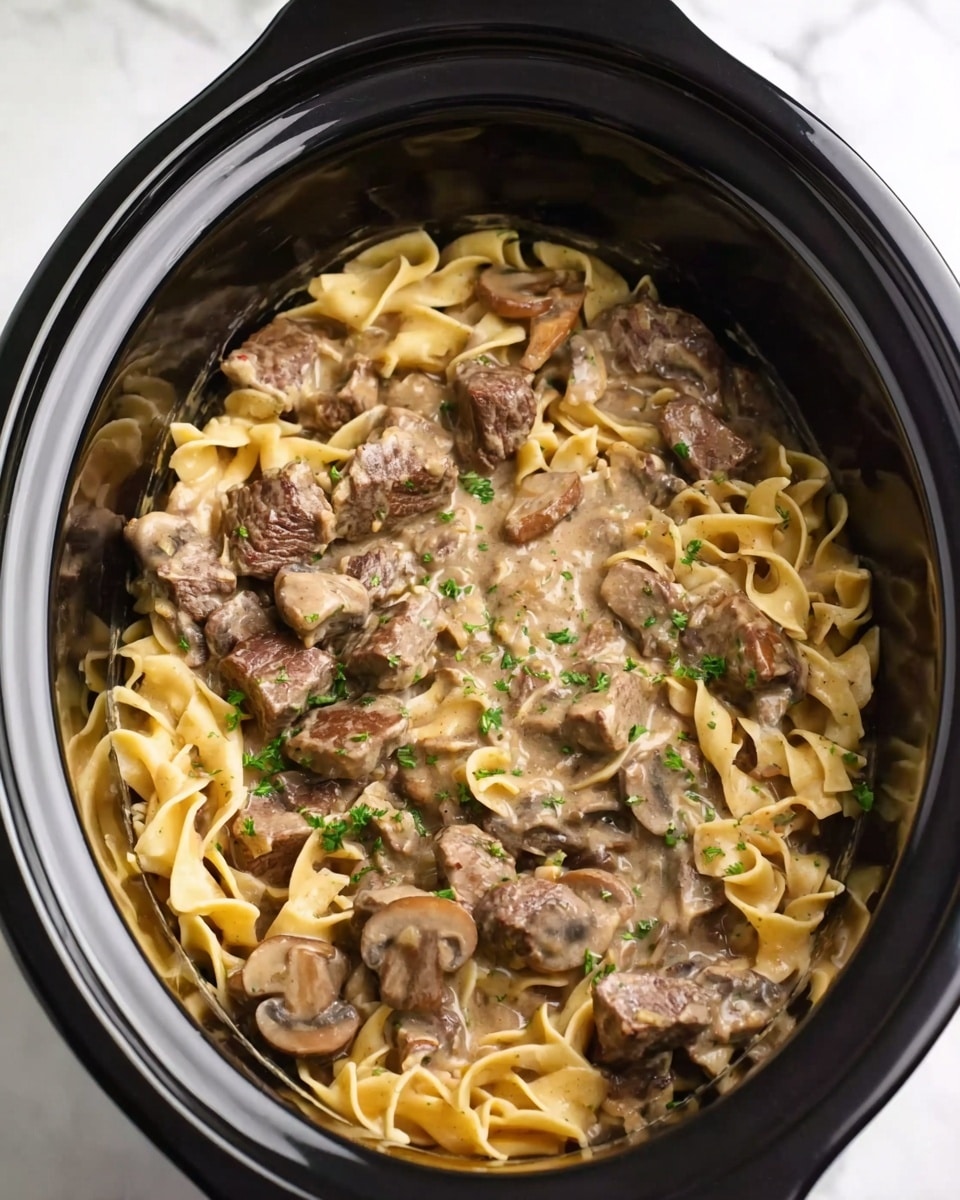 The image shows a slow cooker filled with a creamy beef stroganoff dish. The bottom layer is light yellow egg noodles with a soft texture, mixed evenly throughout the pot. On top of and mixed with the noodles are chunks of browned beef pieces, which are dark brown and juicy. Sliced mushrooms, tan and smooth, are scattered throughout the dish. The whole mix is coated in a thick, creamy sauce that is light brown in color and slightly shiny. Small green parsley bits sprinkle over the dish to add a touch of color. The slow cooker is black, and it sits on a white marbled surface. photo taken with an iphone --ar 4:5 --v 7