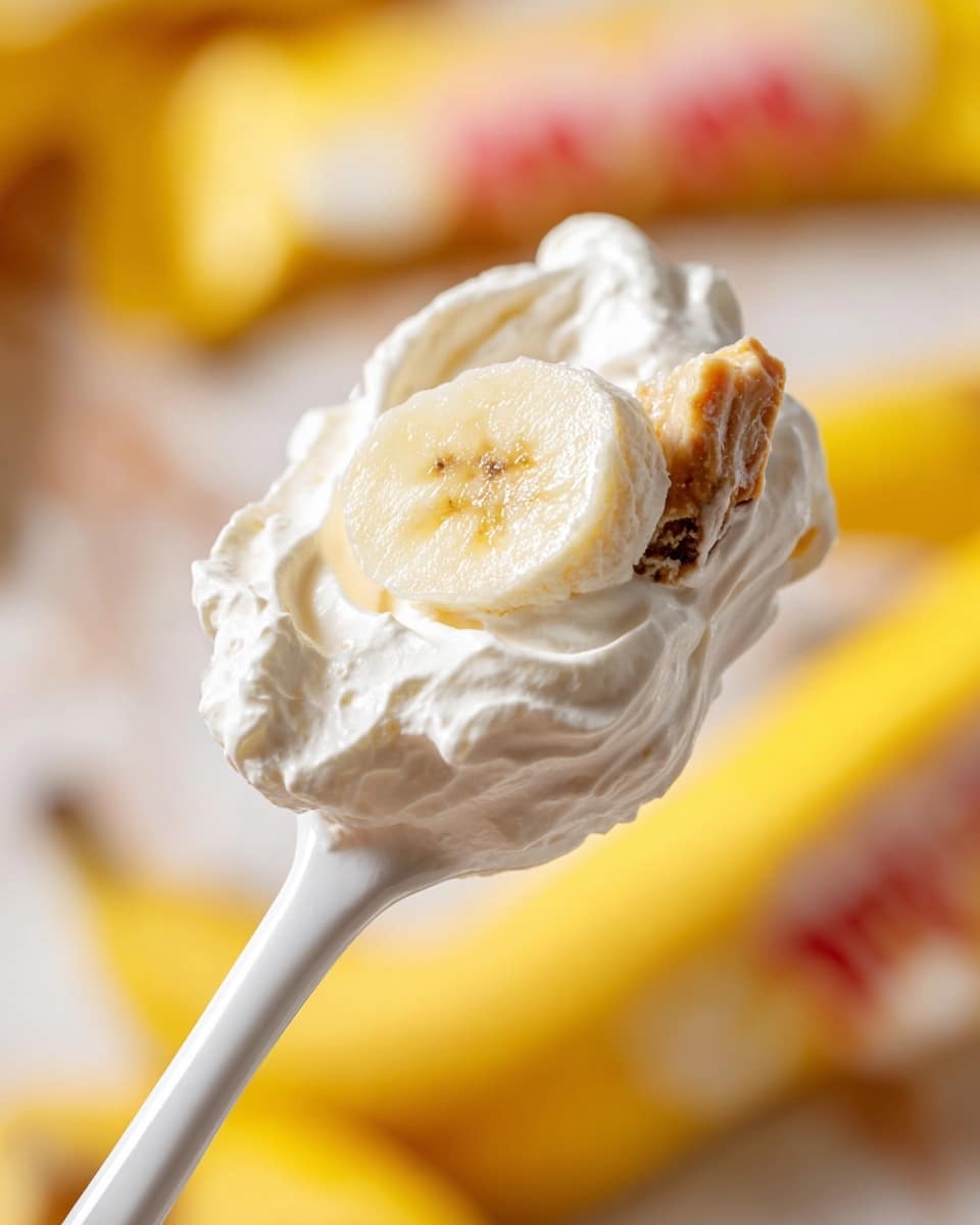 A close-up view of a white spoon holding a dollop of thick, fluffy white cream with a slice of yellow banana and a small piece of a light brown candy bar embedded in the cream, with a blurred background showing parts of a yellow candy wrapper and a white marbled surface. photo taken with an iphone --ar 4:5 --v 7