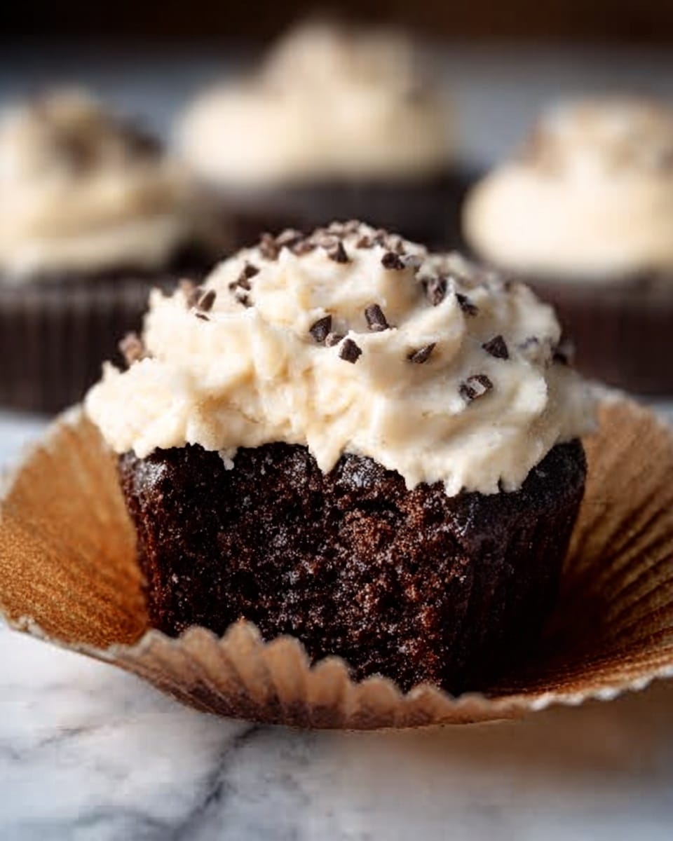 A close-up of a single chocolate cupcake sitting inside an open brown paper liner on a white marbled surface, topped with a thick layer of creamy white frosting mixed with small chocolate pieces, the cupcake's dark and moist texture visible beneath the smooth frosting, with blurred cupcakes in the background. Photo taken with an iphone --ar 4:5 --v 7