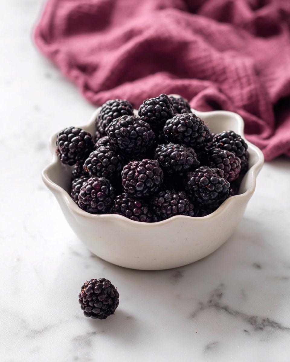 A white ceramic bowl with a wavy edge is filled with shiny, dark purple blackberries, piled high and showing detailed bumpy texture. Two blackberries sit on the white marbled surface near the bowl. In the background, a folded deep pink cloth adds a soft, warm contrast against the clean white marble. The photo is taken with an iphone --ar 4:5 --v 7