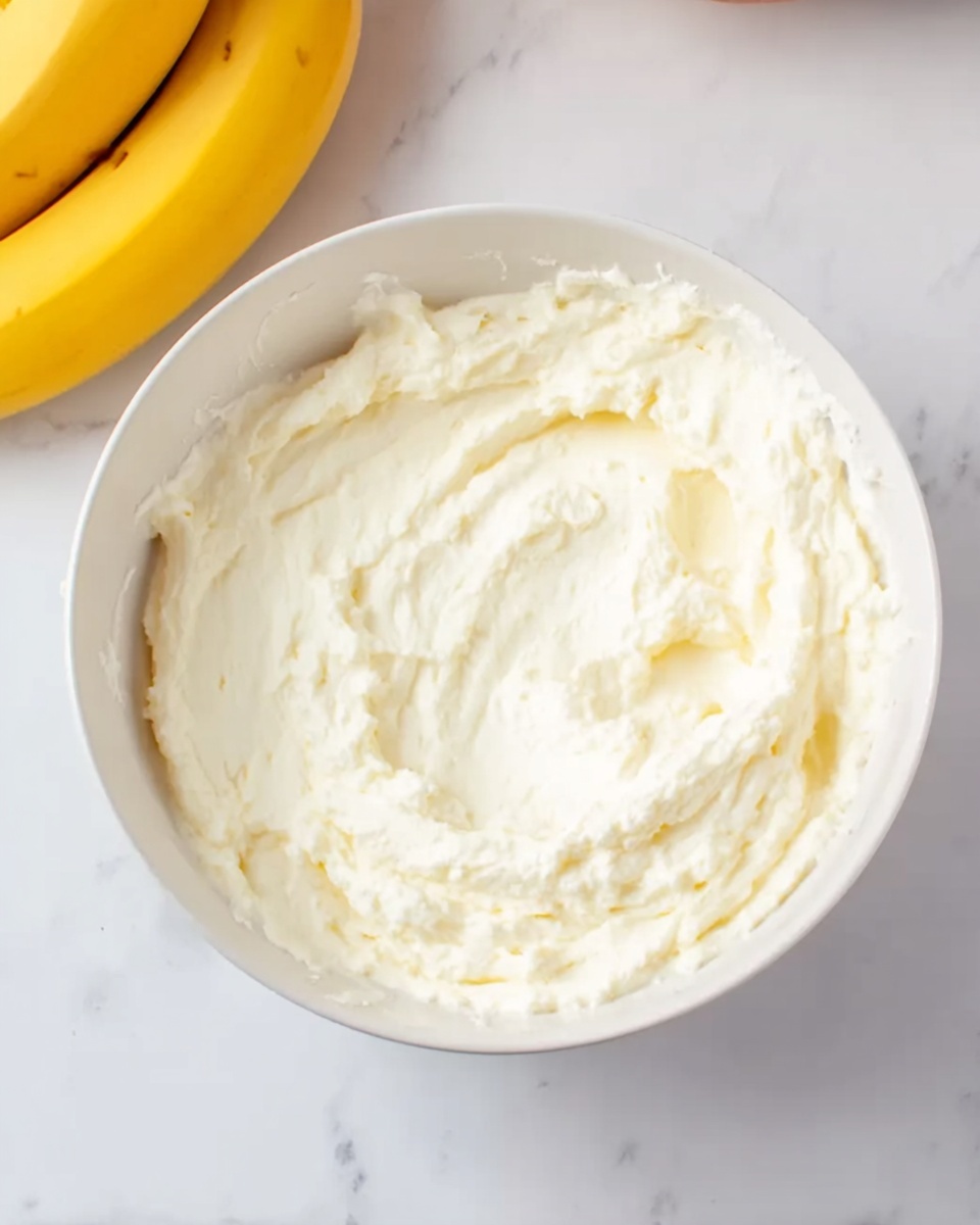 A white bowl filled with a light, creamy mixture that has a soft, fluffy texture, almost like whipped cream or mashed cheese, sits on a white marbled surface. Near the bowl, there are two yellow bananas partially visible on the left side. The bowl and the creamy mixture inside are the main focus, with the smooth white marbled background giving a clean and bright look. Photo taken with an iphone --ar 4:5 --v 7