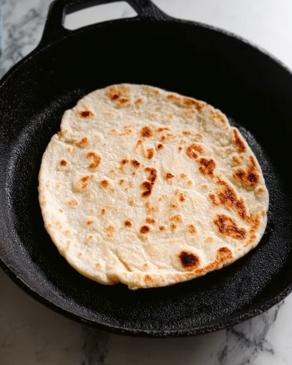 A round, light beige flatbread with some small brown spots is cooking on a black cast iron skillet. The flatbread is slightly puffy in some areas and has a soft texture. The skillet has a matte finish and sits on a white marbled surface. Photo taken with an iphone --ar 4:5 --v 7