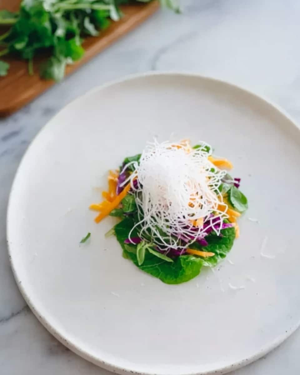 A white round plate holds a small stack of fresh green leaves at the base, topped with thin shredded white noodles arranged loosely. Underneath the noodles, there are small pieces of orange and purple vegetables, with some green herb leaves peeking out from the sides. The plate sits on a white marbled surface with some green herbs off to the upper left side. The colors of the vegetables and noodles create a light, fresh feel in the simple presentation. photo taken with an iphone --ar 4:5 --v 7
