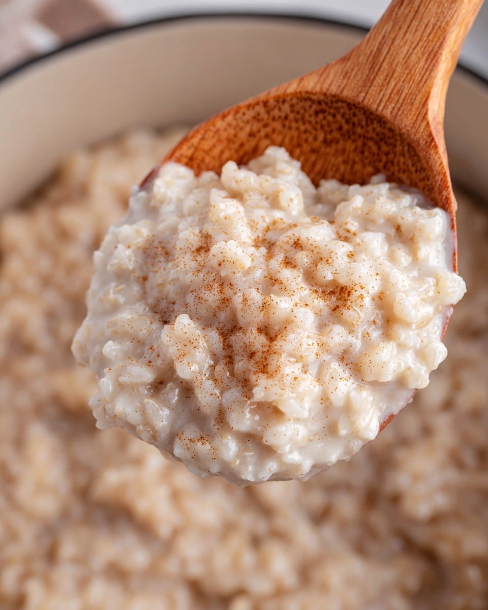 A close-up view of creamy, cooked rice pudding being held by a wooden spoon. The rice pudding shows a soft, slightly grainy texture with a light beige color mixed with tiny brown specks of cinnamon or spices throughout. The background is blurred but suggests more rice pudding in a pot. The image has a warm and cozy feel with the wooden spoon adding a natural touch. Photo taken with an iphone --ar 4:5 --v 7