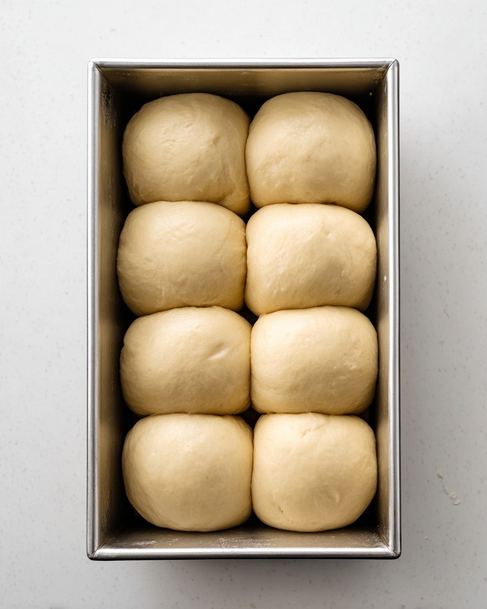 A rectangular silver baking pan holds eight smooth, round dough balls placed closely in two rows of four. The dough balls are light beige with a soft, slightly shiny texture, and they fill the pan almost edge to edge. Each ball is puffy, evenly sized, and touching its neighbors. The pan sits on a white marbled surface, viewed from above in bright, natural light. photo taken with an iphone --ar 4:5 --v 7