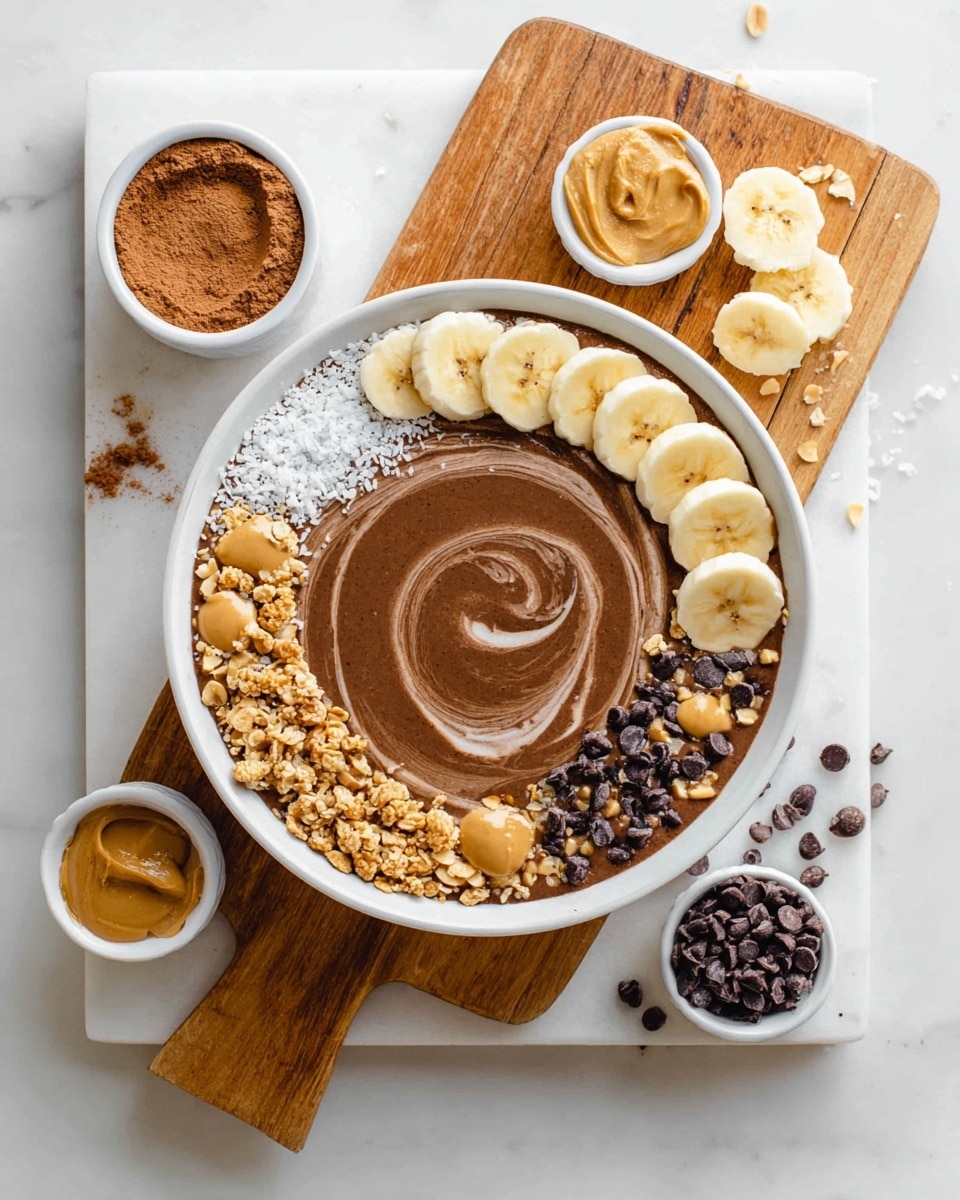 A white bowl filled with smooth chocolate-colored smoothie swirled in the center, topped with arranged layers around the edge including white coconut flakes, light yellow banana slices with small dark specks, light tan peanut butter, light brown peanut pieces, dark chocolate chips, and golden brown granola. The bowl sits on a white marbled board with a wooden cutting board on the top right holding more banana slices and crushed peanuts. Around the bowl on the marble are three small white bowls—one with creamy peanut butter, one with cocoa powder, and one with milk. Scattered dark chocolate chips and crushed peanuts add texture across the surface. Photo taken with an iphone --ar 4:5 --v 7