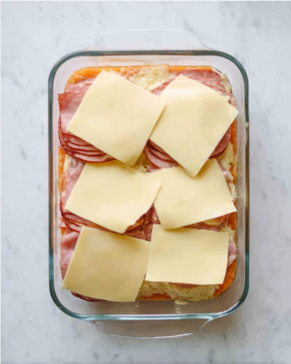 A glass baking dish sits on a white marbled surface, filled with a layered dish. The bottom visible layer is soft orange, likely cooked sweet potatoes or squash, slightly peeking out at the edges. Above this is a layer of thinly folded pink meat that covers the entire dish evenly. On top are four slices of pale yellow cheese, two on the top row and two on the bottom row, with the bottom left slice cut in half and spaced apart slightly. The cheese covers most of the dish but some meat and orange color show around the edges. Photo taken with an iphone --ar 4:5 --v 7