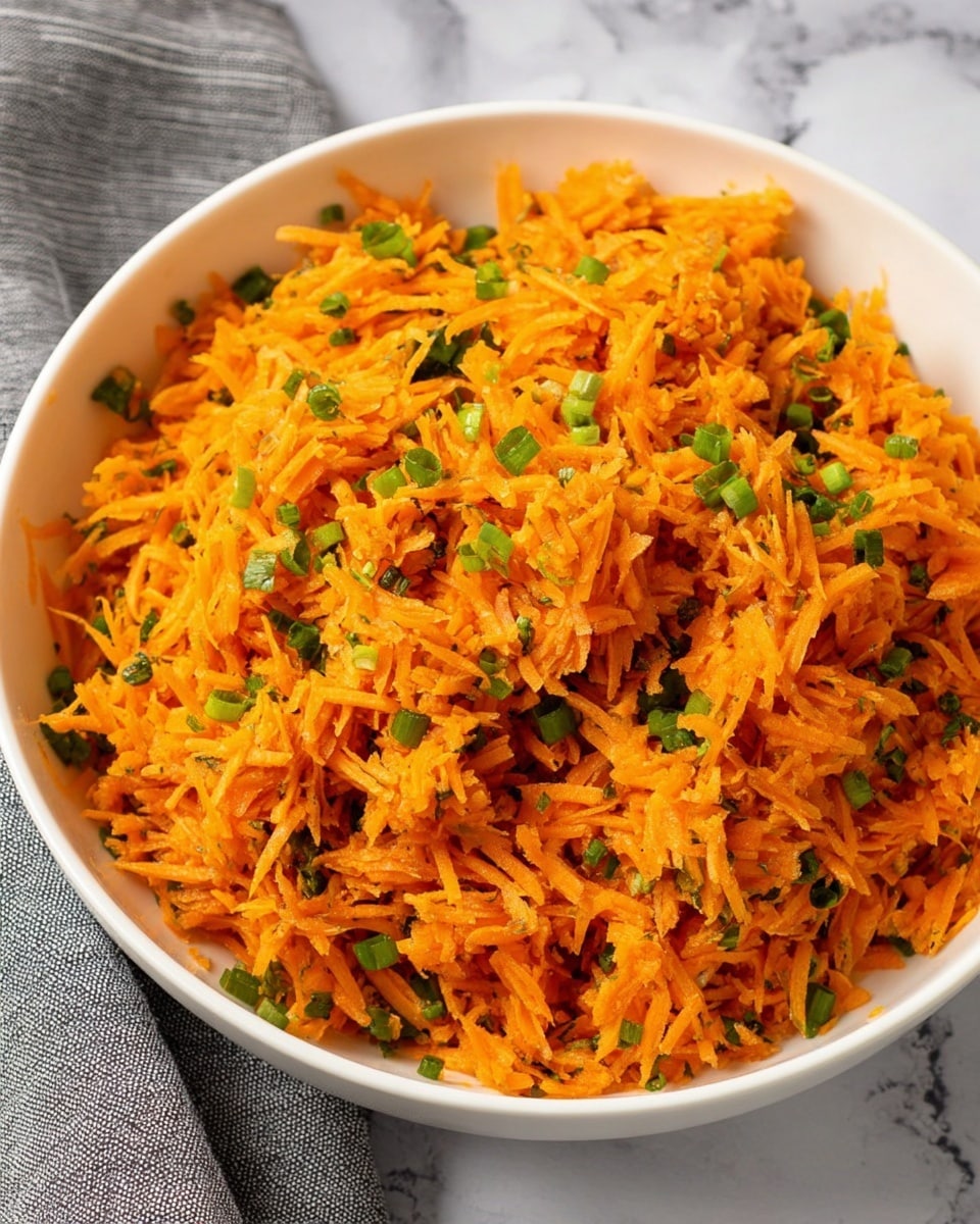 A white bowl filled with finely shredded orange carrots mixed with small green pieces of chopped scallions and herbs evenly spread throughout. The texture looks soft but slightly rough from the shredded carrots, with vibrant orange and green colors contrasting. The bowl rests on a white marbled surface with part of a grey cloth napkin seen in the background. photo taken with an iphone --ar 4:5 --v 7