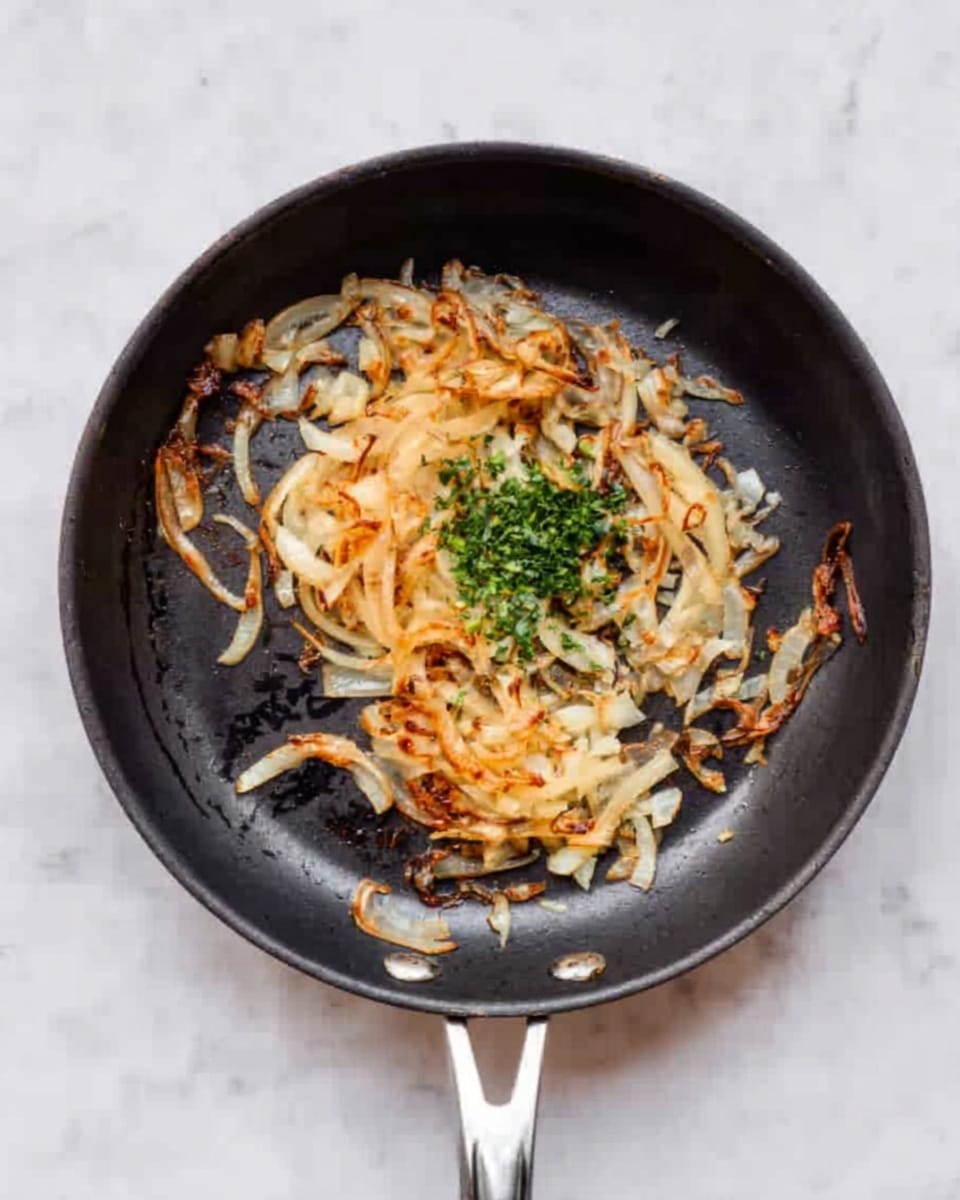 A black frying pan with a silver handle sits on a white marbled surface. Inside the pan, there is a layer of light golden-brown cooked onions, some pieces are slightly crispy and curled. In the center, there is a small pile of chopped green herbs adding a fresh contrast to the warm colors of the onions. The onions are unevenly spread, leaving part of the pan's surface visible. The overall look is rustic and simple, highlighting the cooked onions with a touch of green freshness. photo taken with an iphone --ar 4:5 --v 7