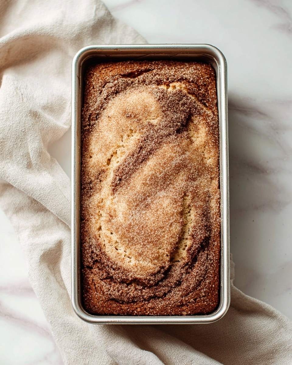 A single-layer loaf cake in a silver rectangular pan sits on a white marbled surface next to a soft, off-white cloth. The cake has a rough, slightly cracked top with a golden brown color and is dusted evenly with a fine layer of light brown cinnamon sugar. The texture looks moist and spongy with darker swirls visible beneath the sugar topping. Photo taken with an iphone --ar 4:5 --v 7