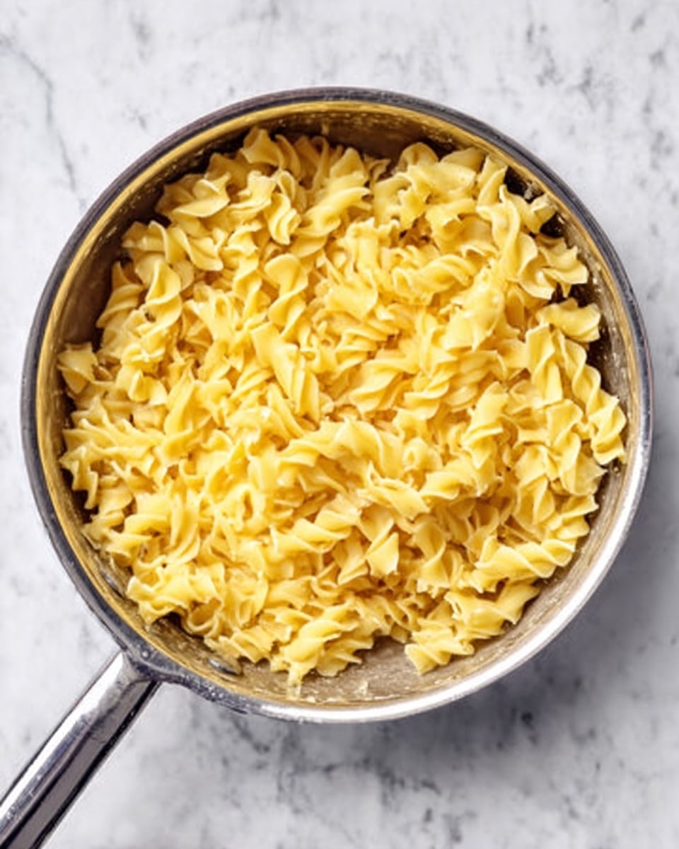 A silver pan filled with curly, yellow cooked pasta, sitting on a white marbled surface. The pasta is evenly spread out inside the pan, showing soft, smooth, and slightly shiny texture. The pan handle extends out to the bottom left corner of the image. The light reflects softly on the pasta and the pan, giving a fresh and clean look. Photo taken with an iphone --ar 4:5 --v 7