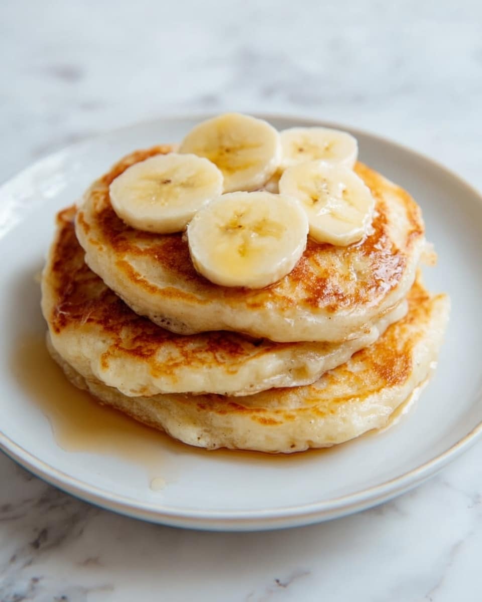 A stack of three golden brown pancakes is placed on a white plate. The pancakes have a soft and fluffy texture with some light browning on the edges. On top of the stack, there are three round slices of banana with a smooth creamy color. The plate rests on a white marbled surface, giving a clean and bright background. photo taken with an iphone --ar 4:5 --v 7