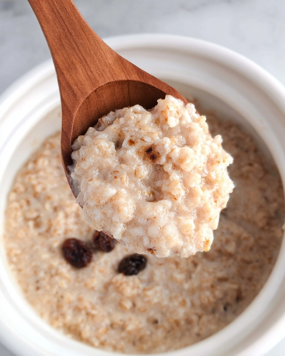 A close-up of a wooden spoon holding a scoop of light brown, creamy rice pudding with visible small grains and a smooth texture. Below the spoon, a white bowl holds more of the same rice pudding, showing a few dark raisins scattered on the surface, sitting on a white marbled texture background. Photo taken with an iphone --ar 4:5 --v 7