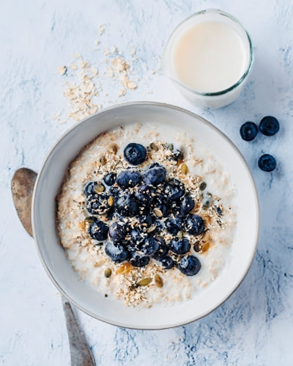 A white bowl filled with creamy oatmeal topped with fresh blueberries and sprinkled with oats and seeds. The blueberries are dark blue and sit in the middle layer over the oatmeal, which is light beige and smooth. The oats and seeds are scattered on top, adding texture with little brown and tan colors. The bowl is placed on a white marbled surface, and there is a small glass of milk and two blueberries nearby. The photo taken with an iphone --ar 4:5 --v 7