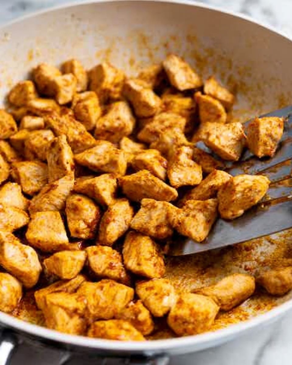 The image shows a close-up of many small, cooked, golden-brown chicken pieces in a white pan. The chicken pieces are cut into small cubes with some showing a light inside texture. A metal spatula is positioned in the pan, lifting some chicken pieces. The pan's surface has a light brown layer of seasoning and cooked bits. The background is a white marbled surface. photo taken with an iphone --ar 4:5 --v 7