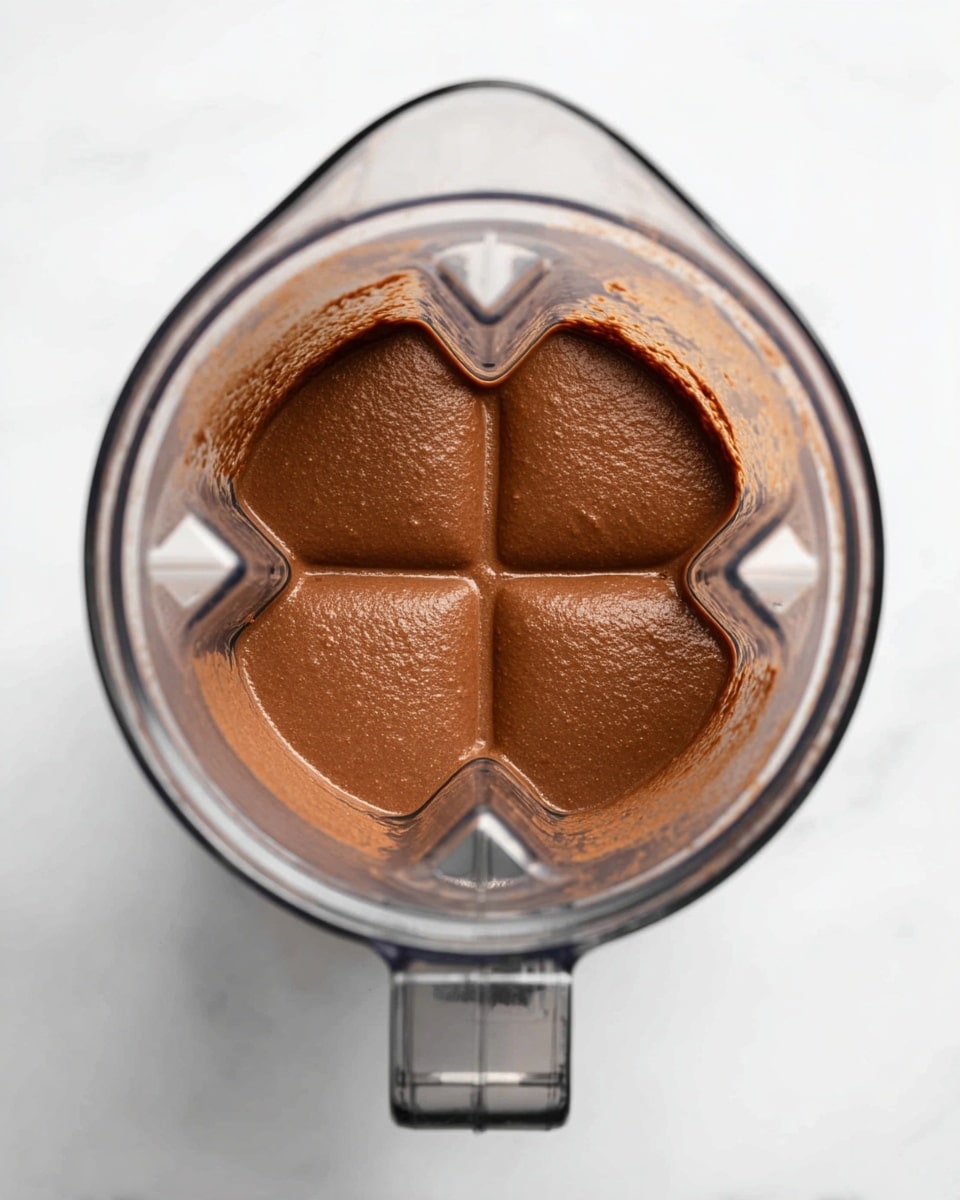 A top view of a clear blender container filled with a thick, smooth chocolate brown mixture, showing four distinct sections where the blades create gentle indentations in the creamy texture. The blender is placed on a white marbled surface, with the handle positioned toward the bottom of the image. The texture of the mixture looks dense and uniform, with a soft shine reflecting light evenly across all areas. photo taken with an iphone --ar 4:5 --v 7