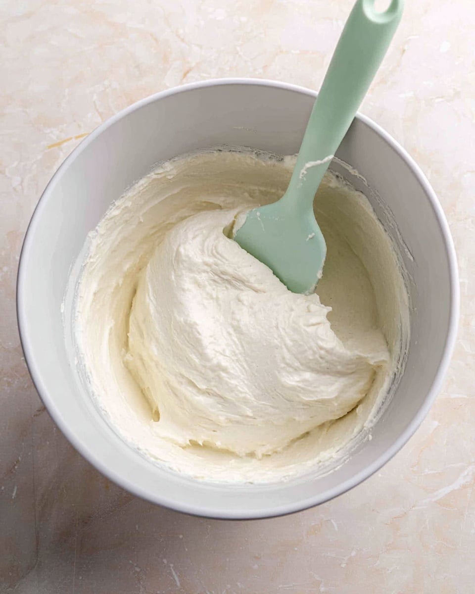 A white mixing bowl filled with a thick, creamy white batter with a smooth, slightly fluffy texture. Inside the bowl, a pale green silicone spatula is partially submerged in the batter, with some batter resting on the spatula’s surface. The bowl is placed on a white marbled surface that adds a clean and soft contrast to the smooth batter inside. Photo taken with an iphone --ar 4:5 --v 7