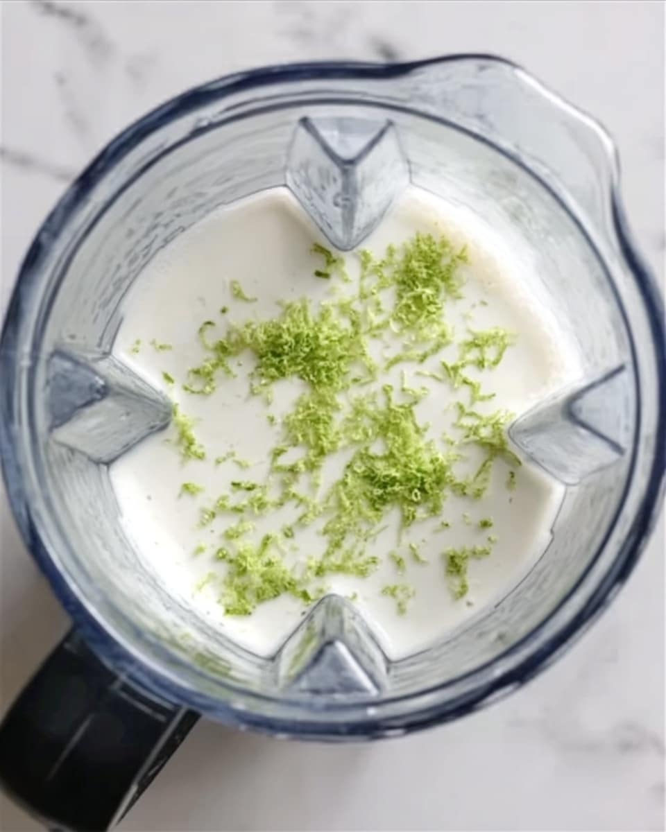 A clear blender container filled with a smooth white creamy mixture at the bottom. On top of the white cream, small bright green lime zest pieces are sprinkled evenly. The blender is placed on a white marbled surface. photo taken with an iphone --ar 4:5 --v 7