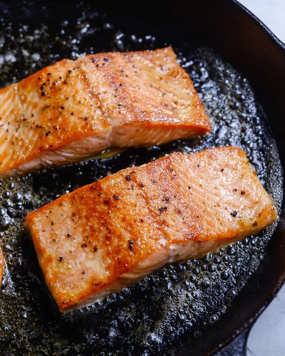 The image shows two pieces of salmon fillets cooking in a black cast iron pan. Each fillet has a golden brown crispy surface with a few black pepper flakes sprinkled on top. The texture looks slightly shiny from the cooking oil, and the salmon skin is not visible from this angle. The pan surface is black and slightly oily with small bubbles around the fish edges, showing it is hot. The background is a clean white marbled texture. photo taken with an iphone --ar 4:5 --v 7