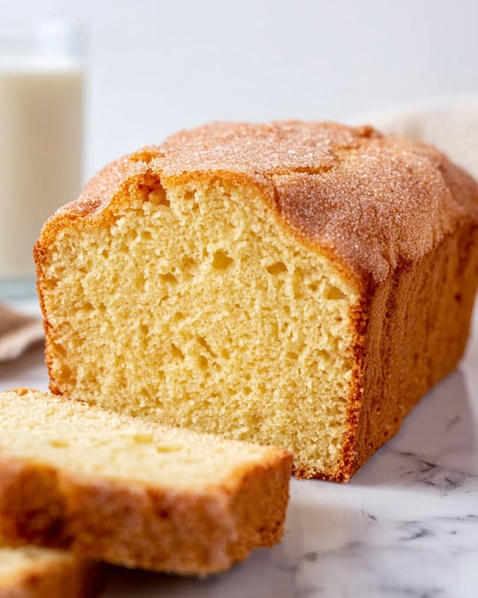 The image shows a thick loaf of light golden yellow cake with a textured, slightly crumbly crust on top. The cake appears soft and moist inside, with small air pockets evenly spread throughout the interior. A slice is shown in front of the loaf, revealing the same light texture. The background is a white marbled surface, and there is a blurred glass of milk visible on the left. The photo taken with an iphone --ar 4:5 --v 7